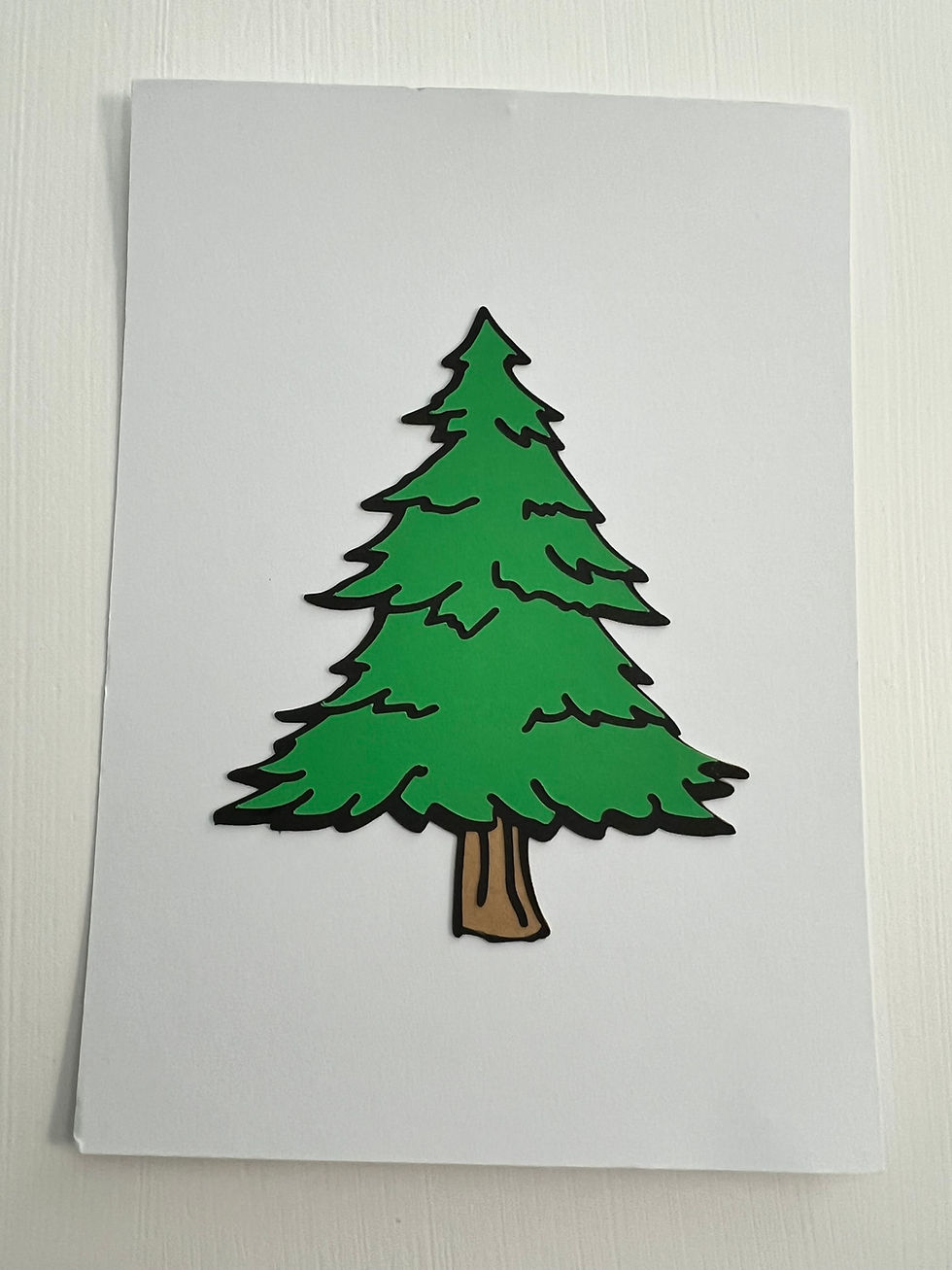 Layered paper evergreen tree with a green top layer, black backing layer, and brown trunk on a white greeting card.