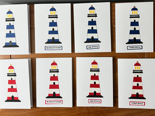 Eight handmade lighthouse cards in red, white, and blue color variations arranged on a wooden surface.