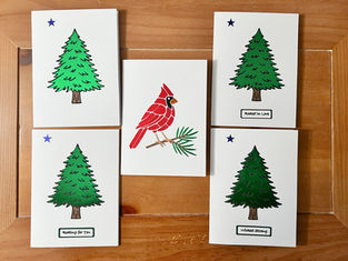 Five handmade cards with pine tree designs and one red cardinal card arranged on a wooden surface.