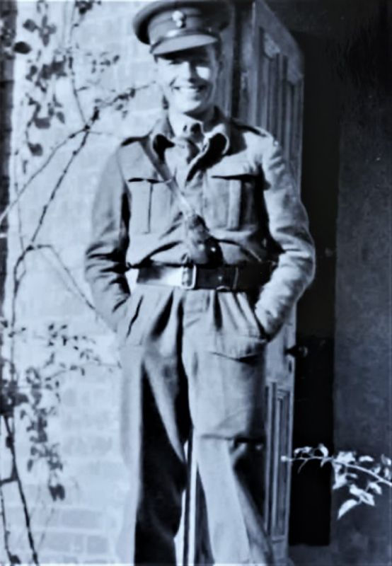 Smiling person in military uniform stands in front of a door with brick and vine background. Vintage-style black and white photo.
