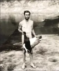 Man standing on rocky terrain, holding a large bird by the legs near a body of water. Black and white photo, rugged landscape, no text.