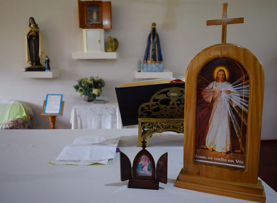 A imagem mostra um altar religioso com várias estátuas e ícones. No centro da imagem, há uma mesa coberta com uma toalha branca. Sobre a mesa, há um ícone de madeira com uma imagem de Jesus e a inscrição "Jesus, eu confio em Vós". À frente deste ícone, há um pequeno triptico com uma imagem religiosa. Ao fundo, há uma mesa menor coberta com uma toalha branca e um arranjo de flores. Na parede, há duas prateleiras com estátuas religiosas, uma delas de Nossa Senhora de Fátima.