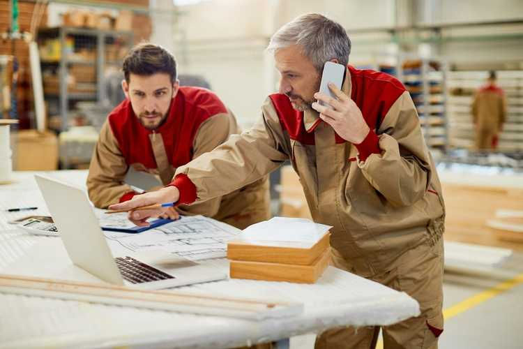 Two factory workers in tan-and-red uniforms review blueprints at a laptop; one points while talking on a phone in a warehouse workshop.