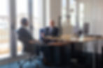 Two men in suits converse in a glass-walled office, with a desk, computer, and city view. Bright setting, professional mood.