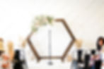 Wooden hexagon arch with floral arrangement, empty mic stand in center. Seated people in floral attire, white geometric wall background. At the Azalea Event Venue in South Jordan, Utah.