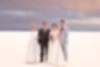 Four people in formal attire stand on a white landscape under a cloudy sunset. Two women in gowns, two men in suits, with a bouquet.