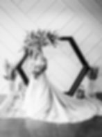 Bride in a flowing gown stands by floral arch, holding a bouquet. Geometric wall, pampas grass, and lanterns in the background. Black and white. At the Azalea Event Venue in South Jordan, Utah.