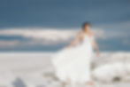 Bride in a flowing white dress stands on a salt flat under a moody sky, looking serene. Background: vast white expanse and dark clouds.