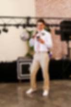 Man in white shirt and beige pants speaks into a mic, reading from phone. Brick wall, disco ball, DJ equipment in background, cheerful mood. At the Azalea Event Venue in South Jordan, Utah.
