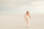 Smiling woman in a flowing dress walks barefoot across a vast, white desert landscape under a cloudy sky, exuding joy and freedom.