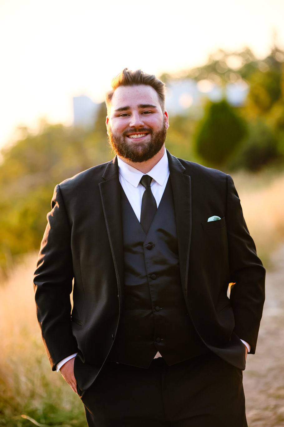 Man smiling in a black suit with a tie, standing outdoors on a sunlit path. Background features soft-focus greenery and warm tones.