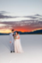 Bride and groom stand on white salt flats, sharing a gaze. Sky glows with orange sunset, mountains in background. Bride holds bouquet.