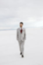 Man in a light gray suit and red tie walking on a vast salt flat, with mountains in the distance under a cloudy sky. Calm demeanor.