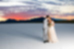 A couple in wedding attire kisses against a vibrant sunset over salt flats, with mountains silhouetted in the background. Romantic and serene.