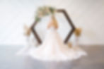 Bride in white gown stands under hexagonal floral arch, white wall background, pampas grass decoration, serene setting. At the Azalea Event Venue in South Jordan, Utah.