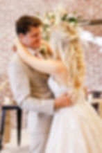 A bride and groom embrace, smiling warmly, against a brick wall adorned with flowers. She wears a tiara, and her dress has lace details.
