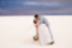 Bride and groom kissing passionately in a desert setting under a dramatic, cloudy sky. Bride holds a bouquet, both appear joyful.