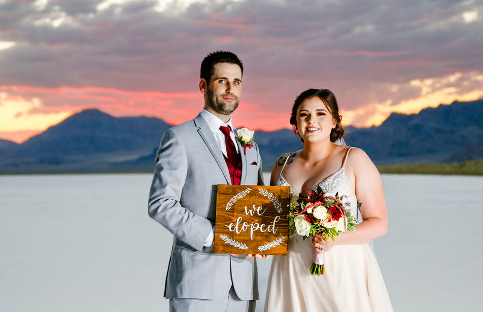 A bride and groom stand together and hole a sign that says, "We Eloped" at the Bonneville Salt Flats with the sun setting behind them.