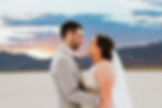Bride and groom embrace at sunset in desert landscape. Bride in detailed gown and veil, groom in light suit. Romantic mood.