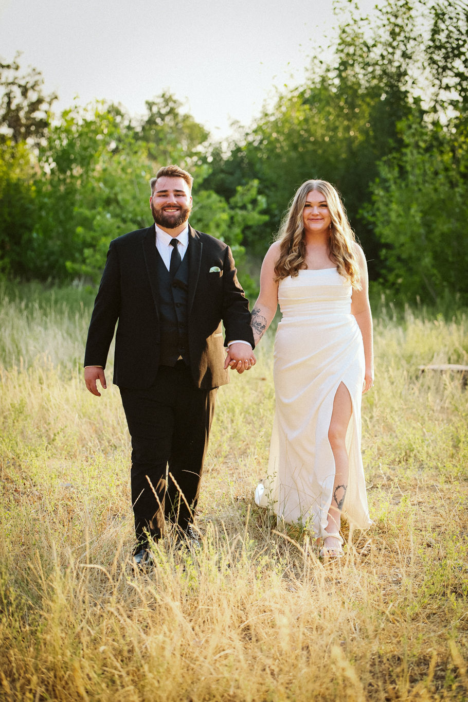A couple holds hands, walking through a sunlit field. He wears a black suit; she wears a white dress with a slit. Green trees in the background.