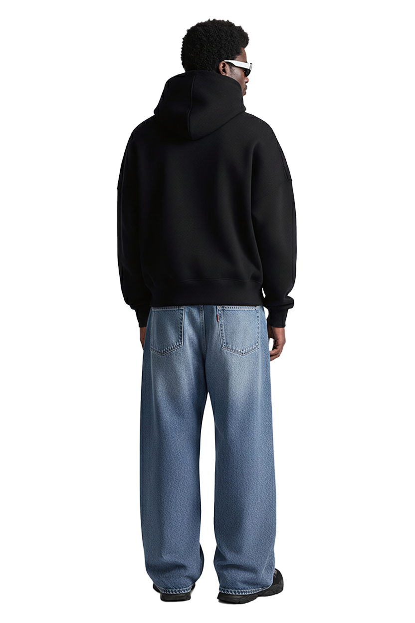 Man in black hoodie and blue jeans, wearing sunglasses, standing against black background.