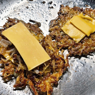 A stainless steel frying pan cooking two hash browns they have cheese and onions on top of them