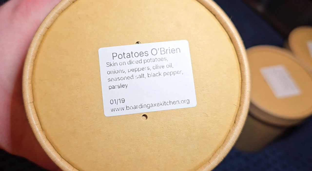 A close up picture of Potatoes O'Brien packaged for the food shelf in a paper cup container. The label on the lid has Title, Ingredients,  Date and Boarding Axe Kitchen on it. 