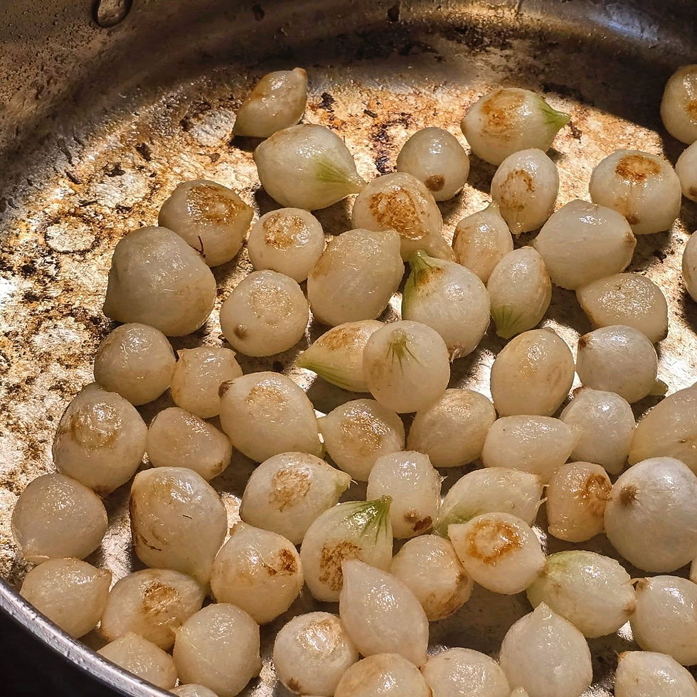 A stainless steel pan filled with white pearl onions that have been partially cooked. They are starting to turn brown and frond can be seen in the bottom of the pan.