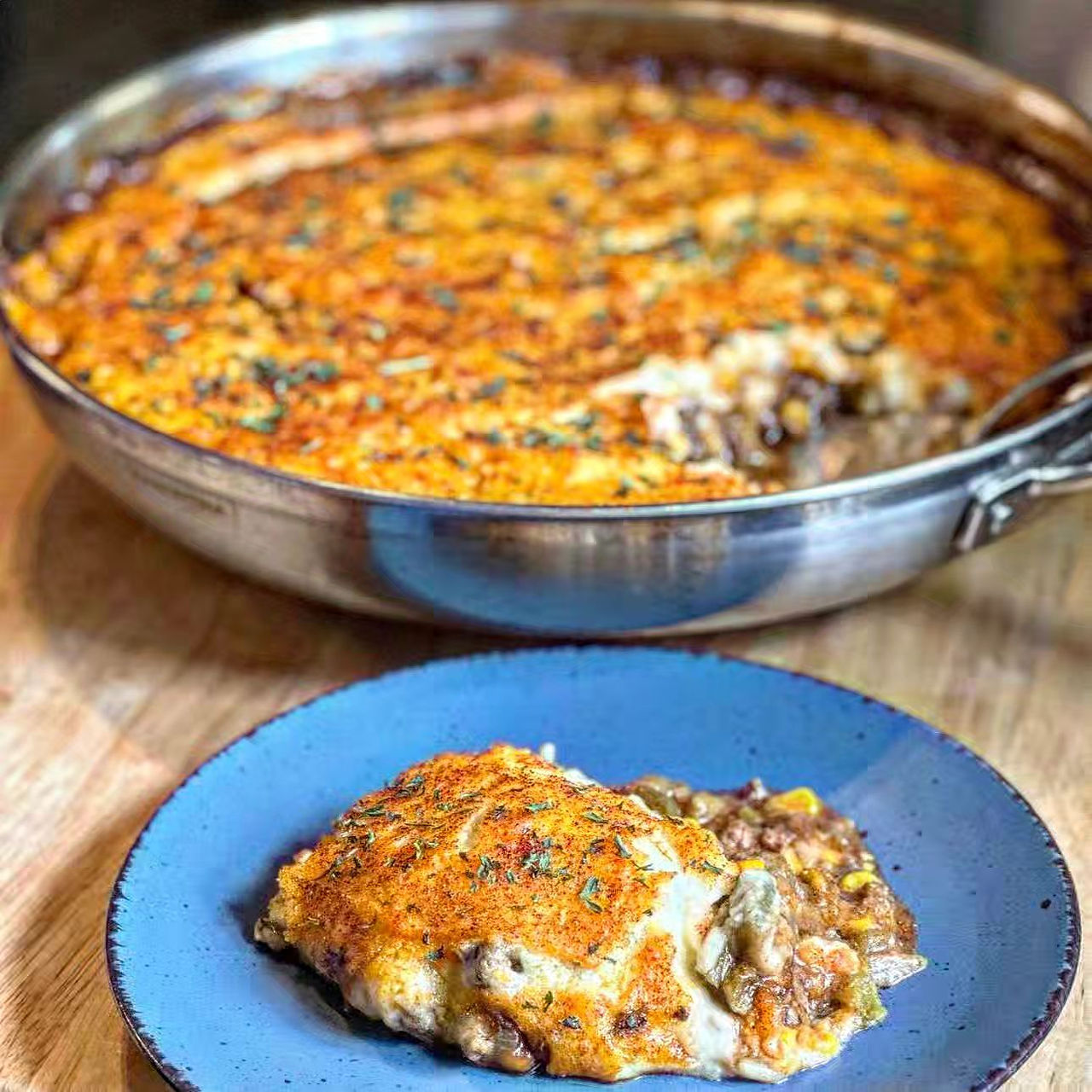 A serving of cottage pie is displayed on a blue dinner plate in front of a stainless steel pan containing the rest of it
