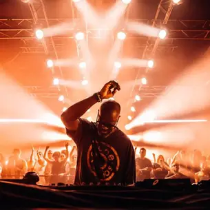 Black Coffee performing an Afro House DJ set in front of a packed club crowd with warm orange stage lights.