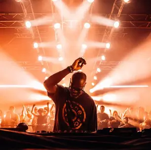 Black Coffee performing an Afro House DJ set in front of a packed club crowd with warm orange stage lights.