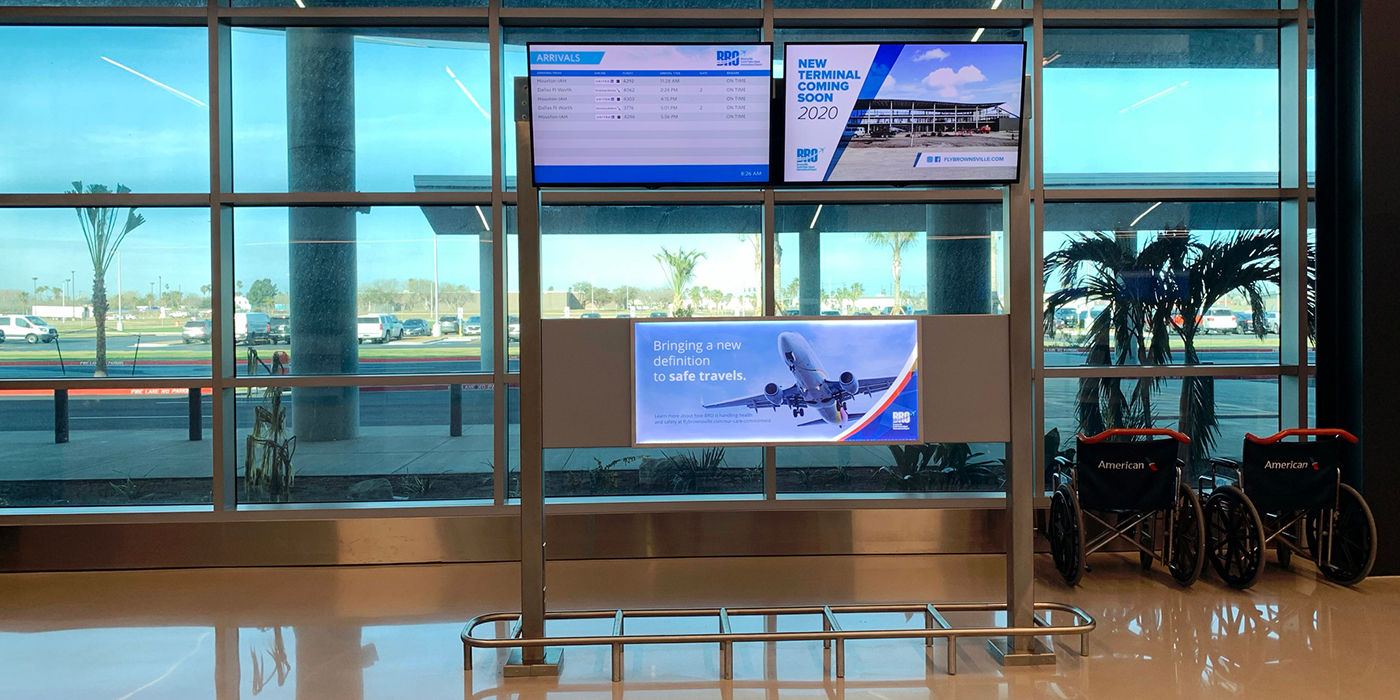 Dual-screen flight and ad display mounted in front of large glass airport windows.