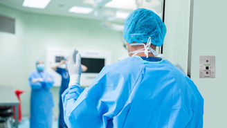 Ein Arzt in einem Krankenhaus trägt einen blauen Kittel und ein blaues Haarnetz und geht durch eine Tür, während er mit der Hand vor einem Parabit-Geste zum Öffnen des Schalters eines berührungslosen Türöffnungsgeräts winkt
