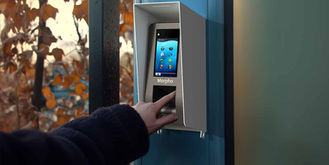 Eine Hand mit einem Finger berührt einen biometrischen Leser mit einem Parabit-Gehäuse, der an einer Wand befestigt ist, hinter der Herbstlaub zu sehen ist
