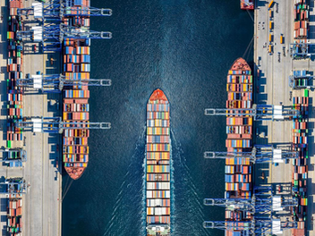Ship passing through shipping port stacked with containers. This represents uncertainty with recent tariffs being placed.