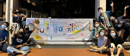 Students in masks sitting around a white banner with the words "Just transition" on it in various colours
