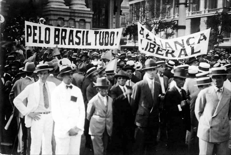 Manifestantes da Aliança Liberal marchando nas ruas em 1929. Em primeiro plano, homens de terno e chapéu, e ao fundo, duas grandes faixas com os dizeres "PELO BRASIL TUDO!" e "ALLIANÇA LIBERAL".