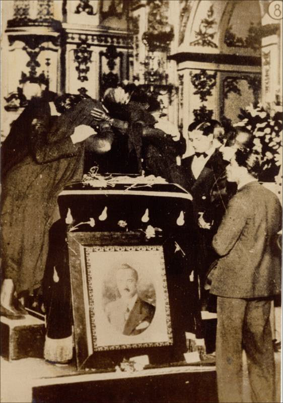 Fotografia em preto e branco do funeral de João Pessoa em 1930. Pessoas se reúnem em luto ao redor do caixão, que está coberto por um pano escuro. Em frente, um retrato emoldurado de João Pessoa.