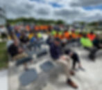 Crowd seated outdoors on a sunny day. People in bright orange and yellow shirts, some standing, others seated. Waiting for speeches to begin.