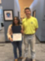 Two people smiling; one holds a certificate. They're indoors with gray carpet, blue walls, and a colorful artwork in the background.