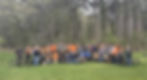 A large group poses outdoors in front of trees, some in orange outfits, on green grass. Two people hold bikes. The mood is cheerful.