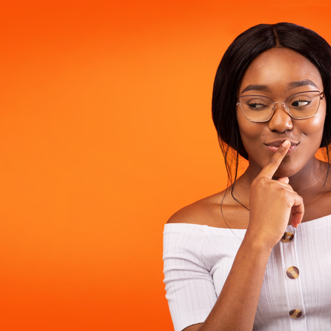 A woman in glasses ponders with a finger on her lips. She's wearing a white off-shoulder top. The background is bright orange.