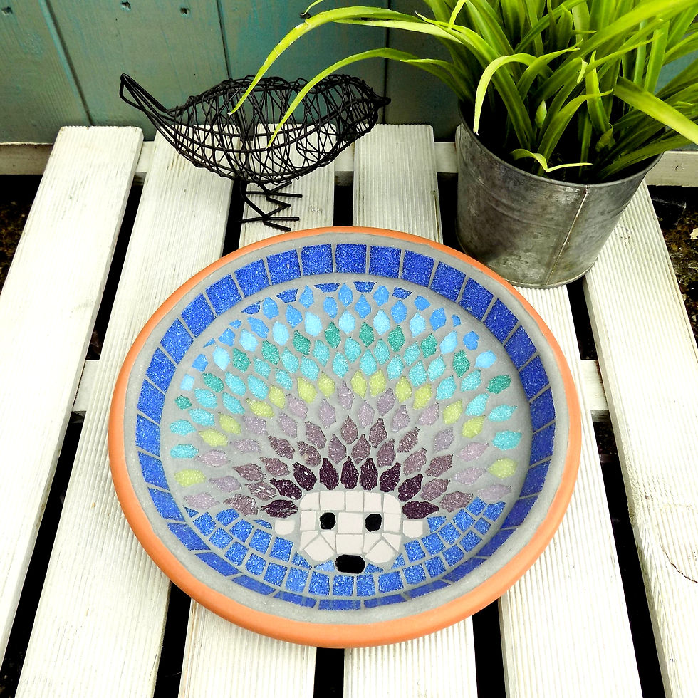 A handmade mosaic garden bird bath with a hedgehog design with spines using shades of greens, purples and blue glass tiles.