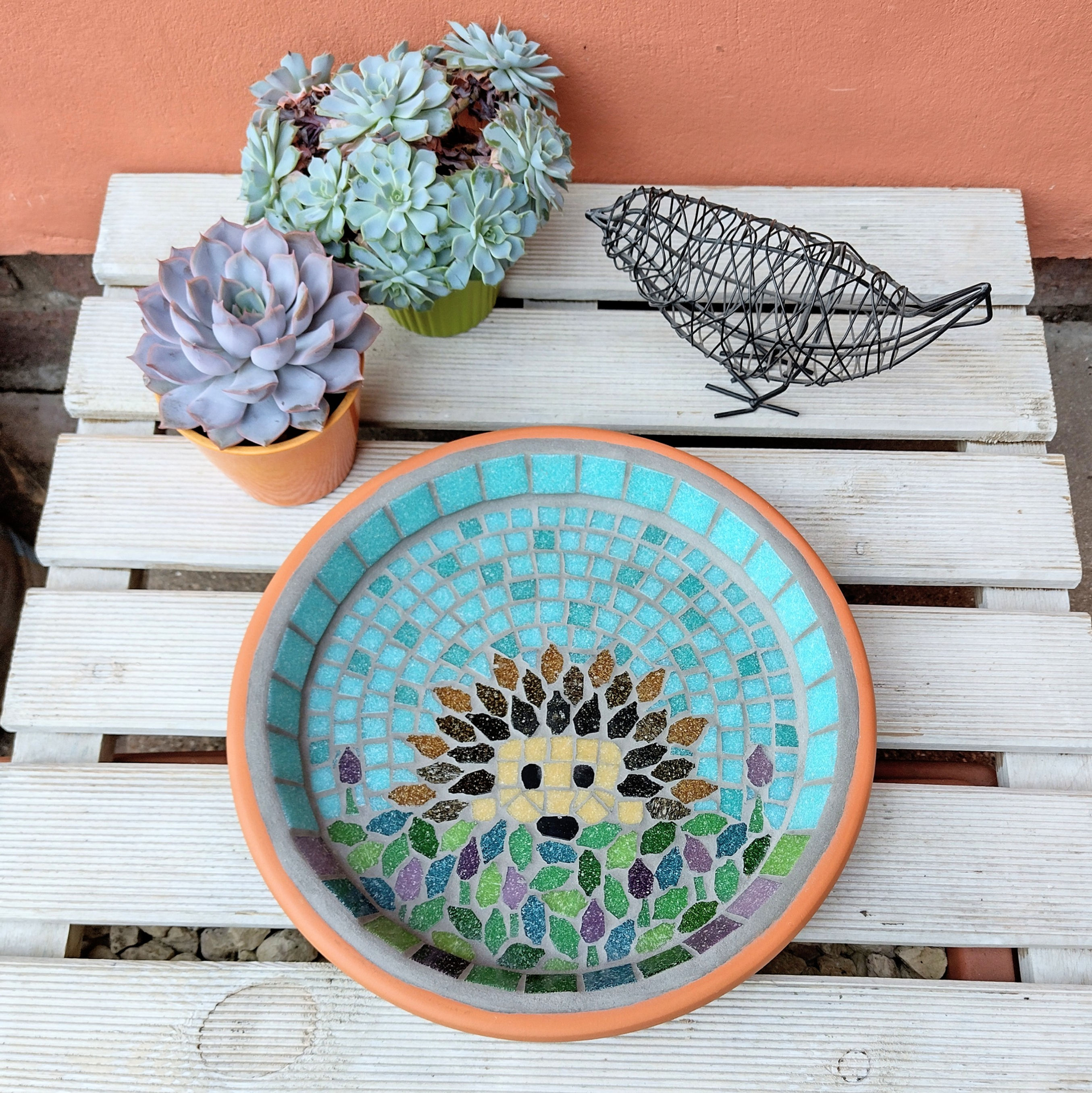 A garden bird bath with a mosaic design of a hedgehog on an abstract lavender flower covered lawn, against a sea green sky.