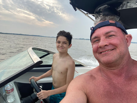 Smiling man and shirtless boy ride a speedboat on a lake at dusk, with cloudy sky and water spray behind them.