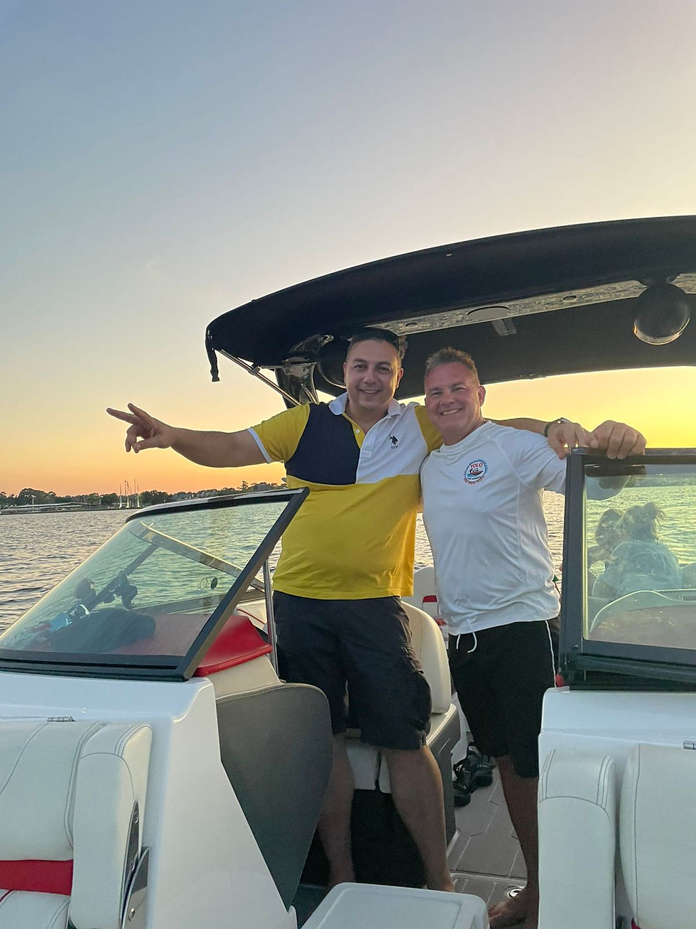 Two smiling men pose on a motorboat at sunset, one flashing a peace sign, with calm water and a glowing sky.