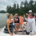 Group on a boat toast with champagne on a calm lake, surrounded by trees; one shirt reads HUK.