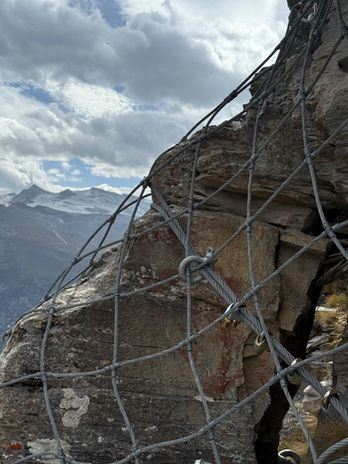 Mit Stahlseilen und Netz gesicherter Felsblock vor dem Bergpanorama.