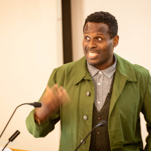 Photograph depicting Dr Onyeka Nubia speaking at a microphone in an animated fashion, wearing a green jacket.