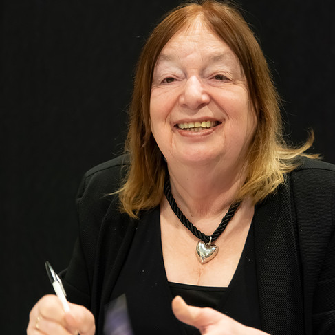 Photograph depicting Dr Alison Weir smiling into the camera while holding a pen. Her clothing is dark against the background.